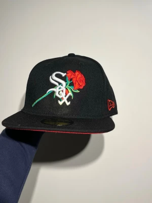 Chicago New Era  59FIFTY keps - Säljer en svart New Era 59FIFTY keps med broderad White Sox-logga och röd ros på framsidan. Sidan har ett patch med 'White Sox 95 Years 1901-1995' och baksidan har röd MLB-logga. Undersidan av skärmen är röd för extra kontrast.