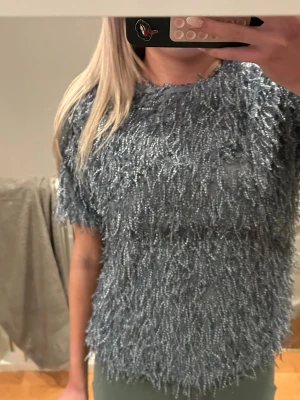 Glittrig blå topp med fransar - Säljer en unik  blå topp med korta ärmar och massor av fransar som ger en cool textur. Toppen har rund hals och är croppad i modellen. Perfekt för dig som vill sticka ut med en trendig look. Storleken är 164/170 så skulle säga att det passar S