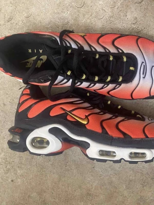 Nike Air Max Plus TN orange och svart - Säljer ett par Nike Air Max Plus TN med grymt färgstark orange och svart ovandel, vita detaljer och guldiga swoosh-loggor. Skorna har den klassiska vågiga designen, synliga Air-enheter i sulan och svarta snören med gula detaljer. Perfekta för dig som vill sticka ut.