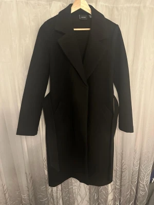Svart kappa med bälte från Vero Moda - Svart lång kappa från Vero Moda med klassisk krage och bälte i midjan och två djupa sidofickor. Perfekt för dig som vill ha en stilren och tidlös look under höst och vinter. Kappan är i vääldigt bra skick  ✨