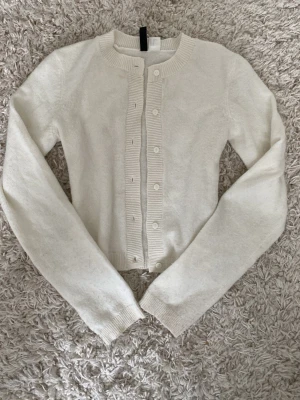 Vit kofta från H&M Divided XS - Säljer en stilren vit kofta från H&M Divided i storlek XS. Koftan har långa ärmar, rund hals och ribbade detaljer vid knappslå och muddar. Den är stickad och har knappar framtill, perfekt för lager-på-lager under kyliga dagar.