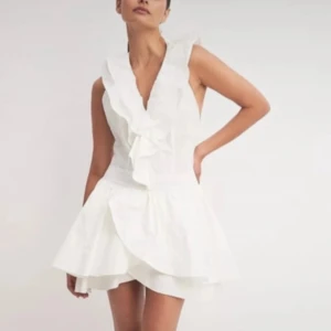 Vit klänning med volang - Supersöt vit kortklänning med omlott-design och volangdetaljer längs halsringningen. Klänningen är ärmlös och har en öppen rygg. Perfekt till sommaren eller student! Har en liiten fläck på innersidan , men syns inte när den är på.