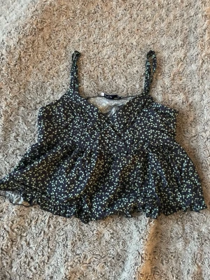 Blommig linnetopp från Hollister XS - Supersöt linnetopp från Hollister i storlek XS. Svart botten med små ljusgröna och vita blommor, v-ringad och med smala axelband. Toppen har volangkant nedtill och är lätt och luftig, perfekt för varma dagar. (Kort i storleken)💐