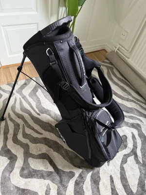 Svart golfbag från Cobra - Snygg svart golfväska från Cobra med sportig design och praktiska detaljer. Västen har flera fack, justerbara remmar och är tillverkad i slitstark polyester med diskret rutmönster. Med ryggkudde samt kylarfack, enbart använd 1 gång
