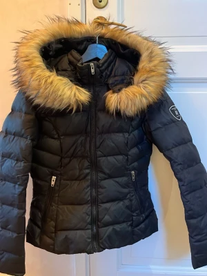 Svart pufferjacka Rockandblue med päls - Svart pufferjacka från Rockandblue med stor fluffig huva och snygg pälsdetalj i beige runt kanten. Jackan har dragkedja framtill och två fickor med dragkedja. Perfekt för kalla dagar och har en cool, modern look.