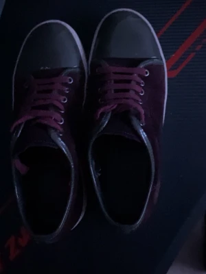 Lanvin vinröda sneakers med mocka - Säljer ett par snygga Lanvin sneakers i vinrött med detaljer i mörkgrått och vit sula. Ovandelen är i mocka med rund tå och snörning i matchande vinröda färg. Klassisk Lanvin-logga under sulan. Perfekt för dig som gillar stilrena och lyxiga sneakers. Min mamma köpte dom åt mig men det blev fel storlek därför säljs dom .