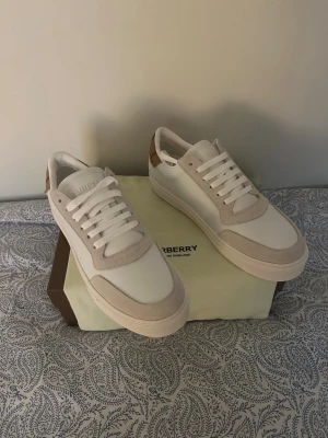 Burberry vita sneakers med beige detaljer - Säljer ett par stilrena Burberry sneakers i vitt med beige mockadetaljer och klassiskt rutigt Burberry-tyg på hälen. Skorna har rund tå, platt sula och snörning framtill. Materialet är en mix av skinn och mocka, med subtila perforeringar på sidan för extra stil.