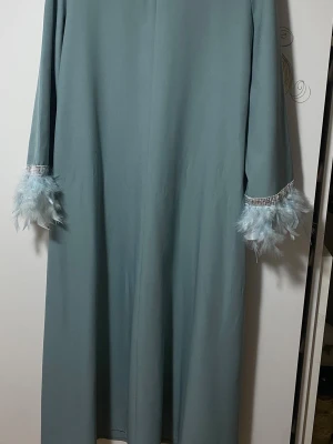  långklänning med fjädrar - Säljer en elegant ljusblå långklänning med raka långa ärmar. Ärmsluten är dekorerade med glittrande band och fluffiga fjädrar i matchande färg. Klänningen har en enkel och stilren design. 