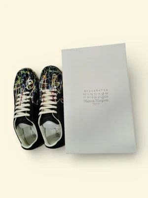 Maison Margiela svarta sneakers med färgstänk - Maison Margiela sneakers i svart skinn med coola färgstänk i vitt, blått, rött och gult. Snygga vita snören och beige gummisula med mönstrad undersida. Klassisk rund tå och platt sula. Kommer med originalkartong.