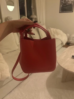 Röd Sanela mini handväska - Säljer en snygg röd mini handväska från Gina  med stilren design och praktiskt axelband. Väskan är tillverkad i slätt syntetmaterial och har ett kompakt format som gör den enkel att bära med sig. Perfekt för dig som gillar minimalistisk look och vill ha en färgklick till outfiten.