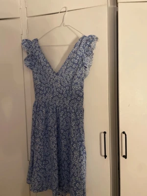 Blommig blå kortklänning med volang - Söt blå klänning med vitt blommönster och volangdetaljer vid axlarna. Klänningen har v-ringad hals och ett knytband i midjan som ger en fin siluett. Perfekt för varma dagar och har en luftig, kort passform.