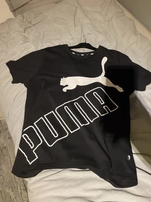 Svart Puma t-shirt med stor logga - Säljer en svart t-shirt från Puma i storlek L med en fet vit Puma-logga och text över hela framsidan. T-shirten har rund hals och korta ärmar, perfekt för dig som gillar sportiga och stilrena plagg. Materialet är mjuk bomull som känns skönt mot huden.