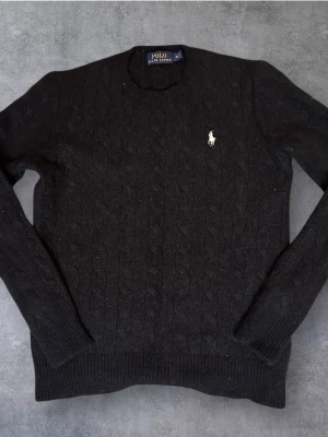 Svart kabelstickad tröja Polo Ralph Lauren - Kabelstickad svart tröja från Polo Ralph Lauren i merinoull. Klassisk rund hals och diskret broderad logga på bröstet. Långärmad modell med ribbade muddar och nederkant. Perfekt för dig som gillar stilrena och lyxiga plagg. Lappen säger storlek M men den har krympt i tvätten så sitter som en XS - S. 38cm bred och 52cm lång. 