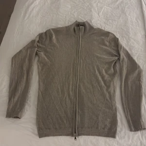 Beige kofta från Oscar Jacobson - Säljer en stilren beige/ljusbrun kofta från Oscar Jacobson i storlek M. Koftan har hel dragkedja framtill, ribbad krage och är långärmad. Materialet är mjukt och känns lyxigt, perfekt för lager-på-lager under hösten och vintern.