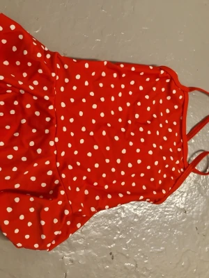 Röd prickig baddräkt med volang - Säljer en röd baddräkt med vita prickar och smala axelband. Baddräkten har en söt volang runt midjan som ger en lekfull touch. Materialet är syntetiskt och passar perfekt för strand eller poolhäng. Supersnygg och färgstark design! Aldrig använt!