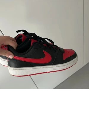 Nike Dunk Low svart/röd sneakers - Säljer ett par Nike Dunk Low sneakers i svart och rött med klassisk röd swoosh på sidan. Skorna har snörning, rund tå och platt vit sula med röd undersida. Ovandelen är i skinn och textil, med ventilerande hål på tån. Perfekt för dig som gillar streetstyle och sportig vibe.