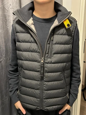 Mörkgrå dunväst från Parajumpers - Snygg svart dunväst från Parajumpers med gul detalj vid kragen och dragkedja framtill. Västen har två sidofickor med dragkedja och en bröstficka. Perfekt för lager-på-lager och har en hög krage för extra värme.