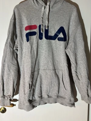 Grå hoodie från FILA med logga - Snygg grå hoodie från FILA med stor logga i blått och rött på bröstet. Klassisk känguruficka framtill och justerbar huva med dragsko. Tillverkad i mjukt sweatshirtmaterial, perfekt för en avslappnad stil.