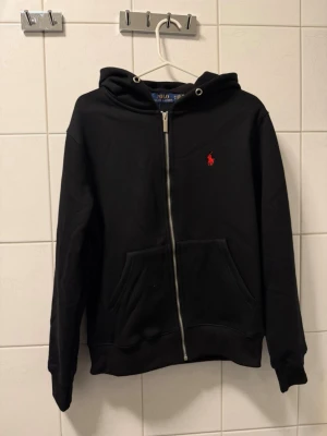 Svart hoodie från Polo Ralph Lauren - Snygg svart hoodie med dragkedja från Polo Ralph Lauren. Klassisk design med röd broderad logga på bröstet, huva och kängurufickor framtill. Tillverkad i mjukt bomullsmaterial som känns skönt mot huden. Perfekt för en avslappnad och stilren look.