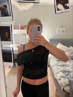 Svart glittrig croppad topp - Snygg svart croppad topp med glittriga paljetter och off shoulder-design. Perfekt för dig som vill sticka ut och glänsa. Toppen har korta ärmar och sitter tight över kroppen. Superfin att matcha med höga jeans eller kjol.
