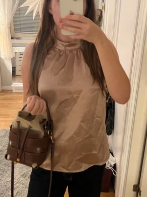 Champagnefärgad ärmlös satinblus - Snygg ärmlös blus i champagnefärgad satin med hög halsringning och lätt glansig finish. Perfekt för dig som gillar en stilren och trendig look. Blusen har en lös passform och är superenkel att matcha med både jeans och kjol.