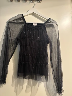 Svart mesh topp med glitter från Zara - Säljer en svart mesh topp från Zara med långa ärmar och glittriga detaljer. Toppen har volanger framtill och är transparent i ärmarna för en extra cool look. Perfekt för dig som vill sticka ut lite extra.