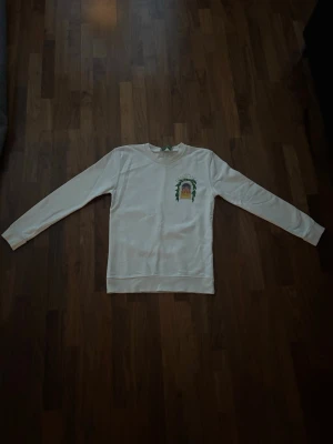 Vit sweatshirt med Casablancatryck - Vit sweatshirt från Casablanca med färgglatt tryck på bröstet och stort emblem på ryggen. Tröjan har rund halsringning och långa ärmar. Perfekt för dig som gillar streetwear och vill sticka ut med en unik design.