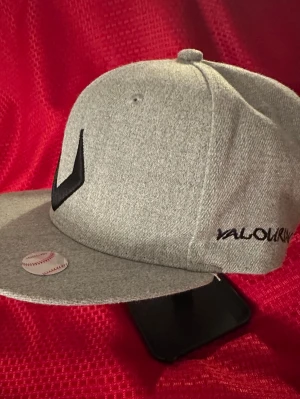 Grå snapback keps från Valourn - Snygg grå snapback keps från Valourn med svart broderad logga framtill och justerbar plastrem bak. Kepsen har rak skärm och är tillverk sopm av ett mjukt tygmaterial. Perfekt för dig som vill ha en clean och modern stil.