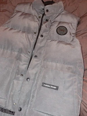 Grå dunväst från Canada Goose - Snygg grå dunväst från Canada Goose med hög krage, två stora framfickor och diskret logga på bröstet. Västen har dragkedja och tryckknappar framtill samt är fodrad för extra värme. Perfekt för lager-på-lager och streetwear-vibe.