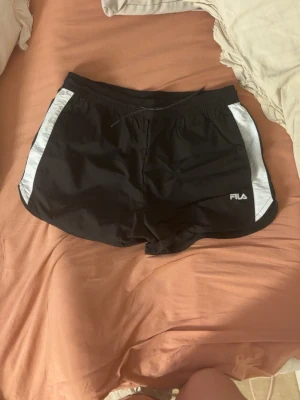 Svarta sportshorts från FILA - Snygga svarta sportshorts från FILA med vita sidopaneler och diskret logga på vänster ben. Shortsen har elastisk midja med snörning för skön passform. Perfekta för träning eller en aktiv dag.