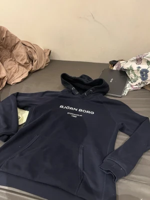 Mörkblå hoodie från Björn Borg - Snygg mörkblå hoodie från Björn Borg med vit text på bröstet där det står 'BJÖRN BORG STOCKHOLM 1956'. Hoodien har en klassisk huva med dragsko och en stor magficka framtill. Perfekt för en avslappnad och sportig stil.