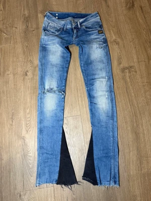 Blå bootcut ”G-STAR” jeans med slits och fransar - Säljer ett par ljusblå bootcut ”G-STAR” jeans med slitningar på knäna och råa fransar vid bensluten. Jeansen har svarta kilar i bensluten för extra vidd och en cool kontrast. Klassisk femficksmodell i jeansmaterial, har funktionerande dragkedjor på båda bakfickorna och är perfekta för dig som gillar en edgy stil.