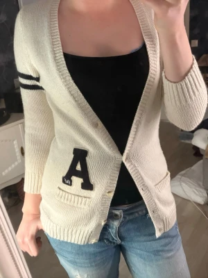 Vit kofta från Abercrombie & Fitch - Vit kofta från Abercrombie & Fitch med svarta ränder på ena ärmen och en stor svart A-patch framtill. Koftan har v-ringning, knappar och två fickor. Stickad i mjukt material och har en klassisk, lite collegeinspirerad look.