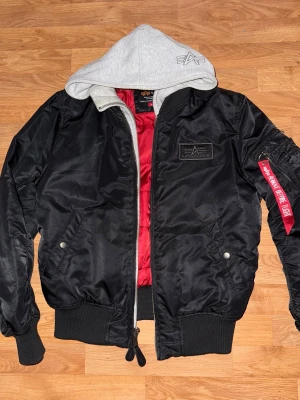 Svart bomberjacka från Alpha Industries - Svart bomberjacka från Alpha Industries med röd innerfoder och grå huva. Jackan har klassisk utility-ficka på ärmen med röd tag, ribbade muddar och dragkedja framtill. Perfekt för dig som vill ha en streetig och cool look.