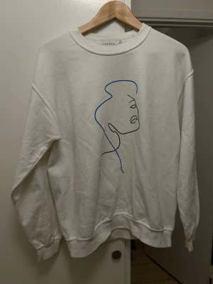 Vit sweatshirt med konstmotiv Topman - Vit sweatshirt från Topman med minimalistiskt konstmotiv i blått och svart på framsidan. Tröjan har rund halsringning, långa ärmar och ribbade muddar. Perfekt för en stilren och modern look.