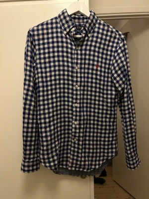 Blårutig skjorta från Polo Ralph Lauren - Klassisk blårutig skjorta från Polo Ralph Lauren med vit bas och blå rutor. Skjortan har lång ärm, knappar framtill och röd broderad logga på bröstet. Perfekt för en stilren och avslappnad look.