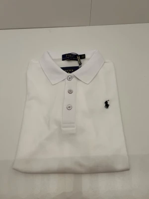 Vit pikétröja från Ralph Lauren - Klassisk vit pikétröja från Ralph Lauren med korta ärmar och tre knappar framtill. Tröjan har den ikoniska broderade loggan i marinblått på bröstet och är gjord i ett mjukt pikématerial. Perfekt till jeans eller shorts för en clean och stilren look. Skriv för annan info