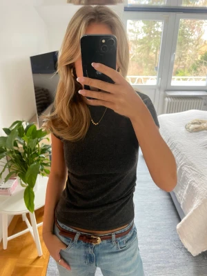 Mörkgrå tight topp med rynk - Snygg mörkgrå topp med tight passform och korta ärmar. Toppen har diskret rynkdetalj i sidan som ger en smickrande siluett. Perfekt att styla med jeans eller kjol! Köpt i Milano