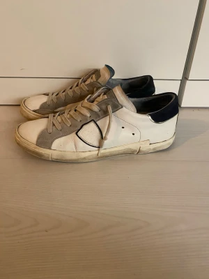 Vita Philippe Model sneakers - Snygga vita sneakers från Philippe Model med grå och mörkblå detaljer. Skorna har klassisk snörning, rund tå och platt sula. Tillverkade i skinn med en stilren design och diskret logga på sidan. Perfekta för en trendig och avslappnad look. Storlek 41 men är väldigt stora i storlek så passar 42