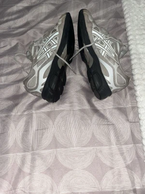 Gråa Asics sneakers med svart sula - Säljer ett par gråa Asics sneakers med klassisk logga i vitt och silver på sidan. Skorna har svart, rejäl sula och snörning framtill. Ovandelen är i mesh och syntetmaterial för bra andningsförmåga och komfort. Perfekta för dig som gillar sportig stil.