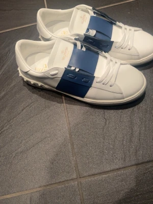 Valentino Open sneakers vit/blå - Snygga Valentino Open sneakers i vitt skinn med bred blå rem över mitten och klassiska nitar på hälen. Skorna har vita snören, rund tå och platt sula. Perfekta för dig som vill ha en clean men ändå unik look. Helt sprillans nya