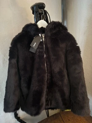 Svart pälsjacka med huva från WEEKDAY  - Mysig svart pälsjacka från Bik Bok med huva och dragkedja framtill. Jackan har en fluffig look och känns riktigt lyxig, perfekt för kalla dagar. Passformen är normal och jackan har långa ärmar. Helt oanvänd med prislappen kvar! 