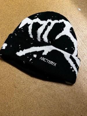 Svartvit mössa från Arc'teryx - Snygg svart mössa från Arc'teryx med vit grafisk design och vikt kant. Tillverkad i mjukt stickat material som håller dig varm och ger en cool streetwear-look. Perfekt för dig som vill ha något unikt och stilrent på huvudet.