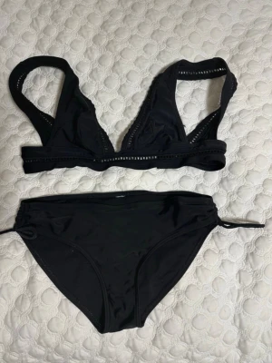Svart bikini med detaljer - Säljer en svart bikini med snygga håldetaljer på toppen och knyt i sidorna på underdelen. Bikinin har en enkel men stilren design och är tillverkad i ett stretchigt syntetmaterial som passar perfekt för strand eller pool. Lite slitet fram på brösten. Storlek S.