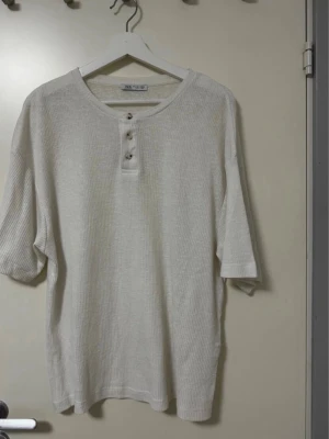 Beige ribbad t-shirt från Zara - Säljer en beige ribbad t-shirt från Zara med tre knappar vid halsen. T-shirten har korta ärmar och en avslappnad passform. Perfekt till jeans eller shorts för en chill look.