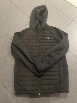 Svart quiltad jacka från Jack & Jones - Säljer en svart quiltad jacka från Jack & Jones med huva och dragkedja framtill. Jackan har släta ärmar, två sidofickor med dragkedja och ett diskret logomärke på bröstet. Perfekt för kyliga dagar och enkel att matcha med allt.