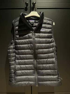 Svart dunväst från Patagonia - Säljer en svart dunväst från Patagonia med quiltad design och dragkedja framtill. Västen har hög krage och är lätt vadderad, perfekt för lager-på-lager. Klassisk och stilren look med diskret logga på bröstet. Har endast använts ytterst få gånger och har en mindre lagning på baksidan. Nypris 2300