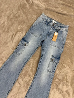 Blå cargopants i jeans med fickor - HELT NYA! ALDRIG ANVÄND! Snygga ljusblå cargopants i jeansmaterial med flera stora fickor på benen och knappdetaljer. Byxorna har en relaxed passform och klassisk cargostil, perfekta för en avslappnad look. Bälteshällor i midjan och cool tvättad finish. stl: 170 