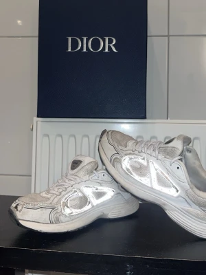 Silvriga Dior sneakers med CD-detaljer - Säljer ett par snygga sneakers från Dior i vitt och silver med reflekterande detaljer och CD-logga på sidan och hälen. Skorna har snörning, rund tå och en chunky sula för extra komfort. Perfekta för dig som vill sticka ut med exklusiva och trendiga sneakers.