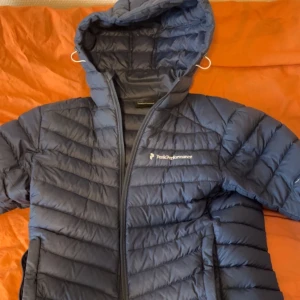 Blå dunjacka Peak Performance - Snygg mörkblå dunjacka från Peak Performance med huva och dragkedja framtill. Jackan har en quiltad design och två fickor med dragkedja. Perfekt för kalla dagar och enkel att matcha med olika outfits.